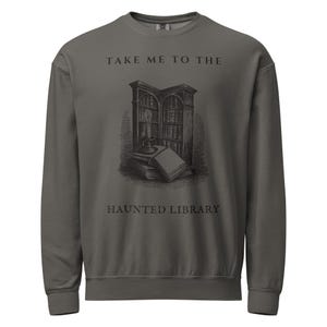 May include: A charcoal grey crewneck sweatshirt featuring a black graphic illustration of a bookshelf, a stack of books, and a lit candle. The text "TAKE ME TO THE HAUNTED LIBRARY" is printed in black capital letters above and below the illustration.