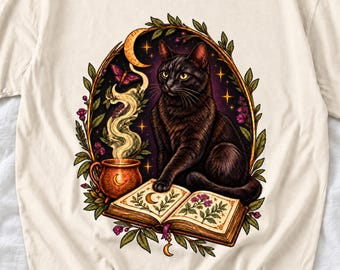 Puede incluir: Una camiseta de color crema con un gráfico de un gato negro sentado sobre un libro abierto. La ilustración incluye un caldero de color cobre humeante, una luna creciente, una mariposa morada y follaje verde enmarcado en un borde ovalado.