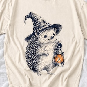 May include: A cream-colored t-shirt featuring a black and white ink-style illustration of a hedgehog wearing a pointed hat. The hedgehog is holding a lantern that emits a warm orange glow, creating a contrast against the monochrome design.