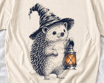 May include: A cream-coloured t-shirt featuring a black and white ink-style illustration of a hedgehog wearing a pointed hat. The hedgehog is holding a lantern that emits a warm orange glow, creating a contrast against the monochrome design.