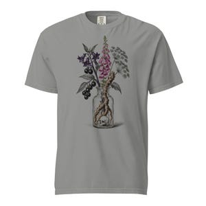May include: A grey short-sleeve t-shirt featuring a graphic print of a clear glass bottle containing a small skull and a root. The bottle is filled with a botanical arrangement of purple foxglove flowers, dark berries, and green foliage.
