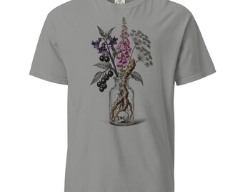 May include: A grey short-sleeve t-shirt featuring a graphic print of a clear glass bottle containing a small skull and a root. The bottle is filled with a botanical arrangement of purple foxglove flowers, dark berries, and green foliage.