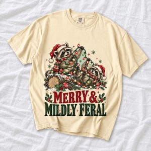 Two raccoons tangled in Christmas lights beside a log with the text Merry & Mildly Feral