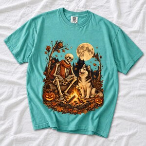 A skeleton in a scarf roasting marshmallows by a campfire beside a husky in an autumn forest under a full moon