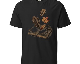 Puede incluir: Una camiseta negra de manga corta con un estampado gráfico de un libro abierto del que emergen varias hojas de otoño marrones y naranjas. El diseño está centrado en el pecho y muestra ilustraciones detalladas de hojas de roble y arce.