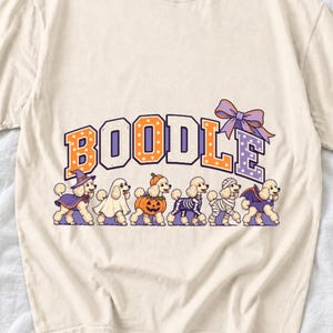 Six poodles in Halloween costumes walking under the arched word BOODLE, lavender and orange polka-dot varsity style