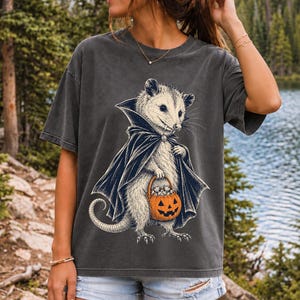 Etched pen illustration of an opossum in a vampire cape holding a candy pumpkin, bone white with orange accent