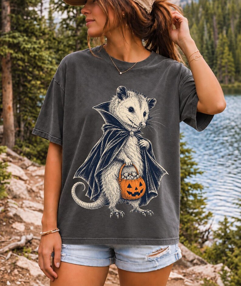 Etched pen illustration of an opossum in a vampire cape holding a candy pumpkin, bone white with orange accent