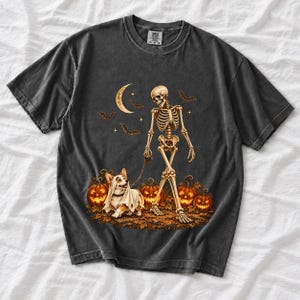 A skeleton walking a corgi in a ghost costume past carved pumpkins under a crescent moon
