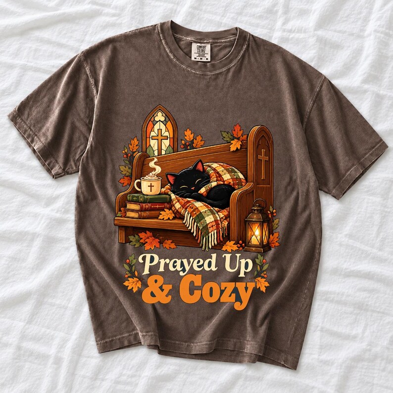 Black cat curled on a church pew with blanket and cocoa among autumn leaves, text "Prayed Up & Cozy", retro illustration