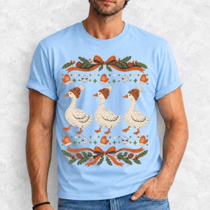 Three geese in pompom knit beanies with mittens, snowflakes and pine borders, retro winter t-shirt graphic