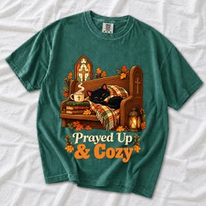 Black cat curled on a church pew with blanket and cocoa among autumn leaves, text "Prayed Up & Cozy", retro illustration