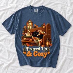 Black cat curled on a church pew with blanket and cocoa among autumn leaves, text "Prayed Up & Cozy", retro illustration