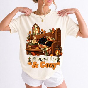 Black cat curled on a church pew with blanket and cocoa among autumn leaves, text "Prayed Up & Cozy", retro illustration