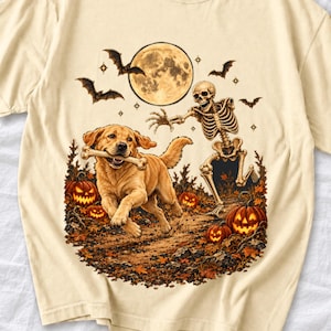 May include: A cream-colored t-shirt featuring a graphic of a golden retriever running with a bone in its mouth, chased by a skeleton. The scene includes a full moon, flying bats, and glowing jack-o'-lanterns amidst autumn leaves.