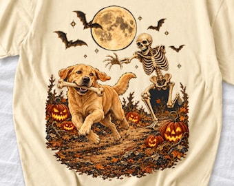 May include: A cream-colored t-shirt featuring a graphic of a golden retriever running with a bone in its mouth, chased by a skeleton. The scene includes a full moon, flying bats, and glowing jack-o'-lanterns amidst autumn leaves.