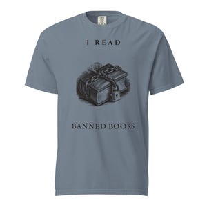 May include: A slate blue short-sleeve t-shirt featuring a black graphic of a book wrapped in chains with a padlock. The text "I READ" is printed above the book, and the text "BANNED BOOKS" is printed below the book in a dark serif font.