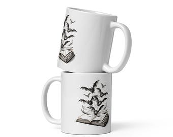 May include: Two white ceramic mugs stacked vertically. Each mug features a black and white illustration of an open book with a swarm of bats flying out from the pages. The design is detailed with ink-style shading and clear, crisp lines.