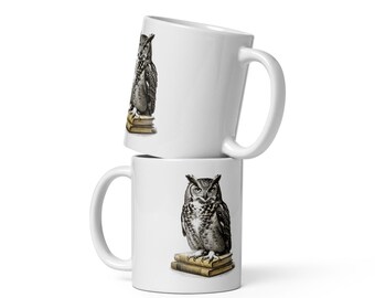 May include: Two white ceramic mugs stacked vertically. Each mug features a detailed black and white illustration of an owl perched on a small stack of tan-colored books. The mugs have a smooth finish, a curved handle on the side, and a cylindrical shape.