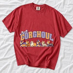 Six corgis in Halloween costumes walking under the arched word CORGHOUL, lavender and orange polka-dot varsity style