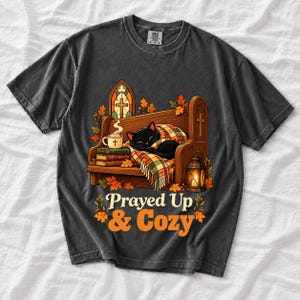 Black cat curled on a church pew with blanket and cocoa among autumn leaves, text "Prayed Up & Cozy", retro illustration