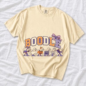 Six poodles in Halloween costumes walking under the arched word BOODLE, lavender and orange polka-dot varsity style