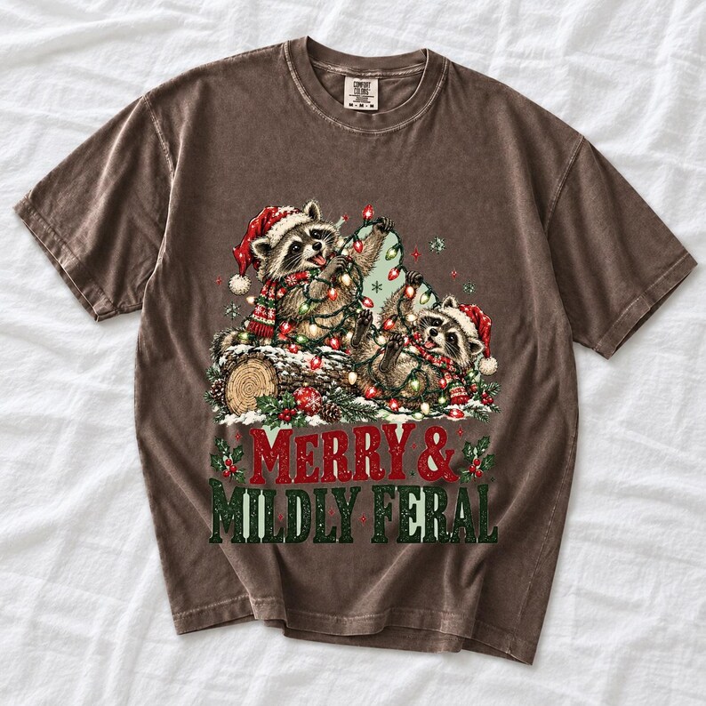 Two raccoons tangled in Christmas lights beside a log with the text Merry & Mildly Feral