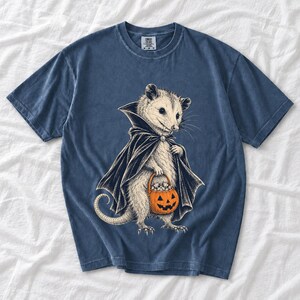Etched pen illustration of an opossum in a vampire cape holding a candy pumpkin, bone white with orange accent