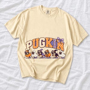 Six pugs in pumpkin, ghost and witch costumes under the arched word PUGKIN, lavender and orange polka-dot varsity style
