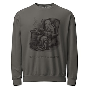 May include: A charcoal grey crewneck sweatshirt featuring a black graphic illustration of a ghost sitting in an armchair reading a book with a steaming cup on a side table. The text below the illustration reads, "TOO COZY TO HAUNT" in black.