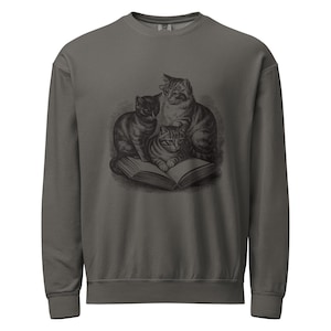 May include: A charcoal grey crewneck sweatshirt featuring a detailed black graphic print of three cats sitting on and around an open book. The long-sleeved garment has a ribbed collar, cuffs, and hem, providing a consistent solid colour throughout.