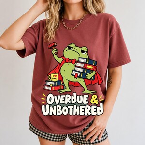 Grumpy cartoon frog librarian holding a stamp and a stack of books with the text Overdue & Unbothered