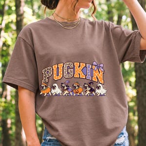 Six pugs in pumpkin, ghost and witch costumes under the arched word PUGKIN, lavender and orange polka-dot varsity style