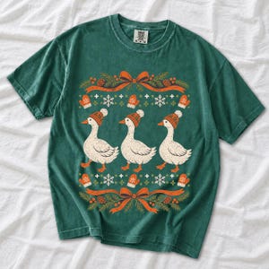 Three geese in pompom knit beanies with mittens, snowflakes and pine borders, retro winter t-shirt graphic