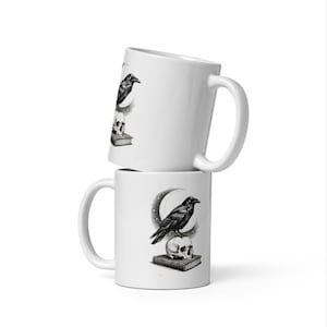 May include: Two white ceramic mugs stacked vertically, each featuring a detailed black and white illustration of a raven perched on a human skull, which rests atop a closed book, all set against a backdrop of a crescent moon and small stars.