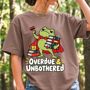 Grumpy cartoon frog librarian holding a stamp and a stack of books with the text Overdue & Unbothered