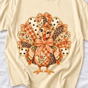 May include: A cream-colored t-shirt featuring a central graphic of a turkey. The turkey has a fan of tail feathers decorated with orange gingham, leopard print, polka dots, and floral patterns, accented with multiple orange bows.