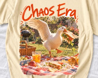 May include: A cream-coloured t-shirt featuring a graphic of a white goose holding a sandwich in its beak. The text "Chaos Era" is printed in red above the goose. The scene depicts a picnic with a basket, a glass of orange juice, and food.