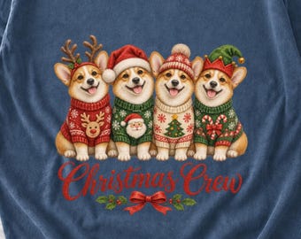 Könnte beinhalten: Ein blaues T-Shirt mit vier Corgi-Hunden in Weihnachtspullovern und Mützen. Die Hunde tragen Rentiergeweihe, eine Weihnachtsmannmütze, eine Strickmütze und eine Elfenmütze. Darunter steht der rote Schriftzug Christmas Crew in Schreibschrift.