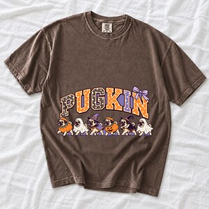 Six pugs in pumpkin, ghost and witch costumes under the arched word PUGKIN, lavender and orange polka-dot varsity style