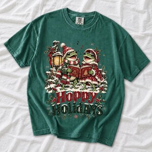 Two frogs singing carols in the snow with a lantern and songbook, text Hoppy Holidays