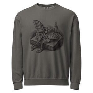 May include: A charcoal grey crewneck sweatshirt featuring a dark graphic print on the chest. The illustration depicts a large moth resting on top of a closed book, positioned next to a single lit candle with a visible flame and dripping wax.