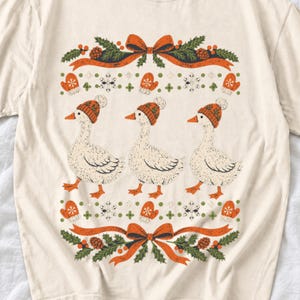 Three geese in pompom knit beanies with mittens, snowflakes and pine borders, retro winter t-shirt graphic