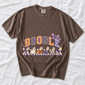 Six poodles in Halloween costumes walking under the arched word BOODLE, lavender and orange polka-dot varsity style