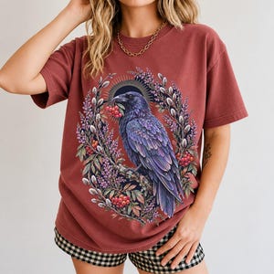 Raven holding a rowan berry inside an oval wreath of heather and catkins, painted cottagecore bird illustration