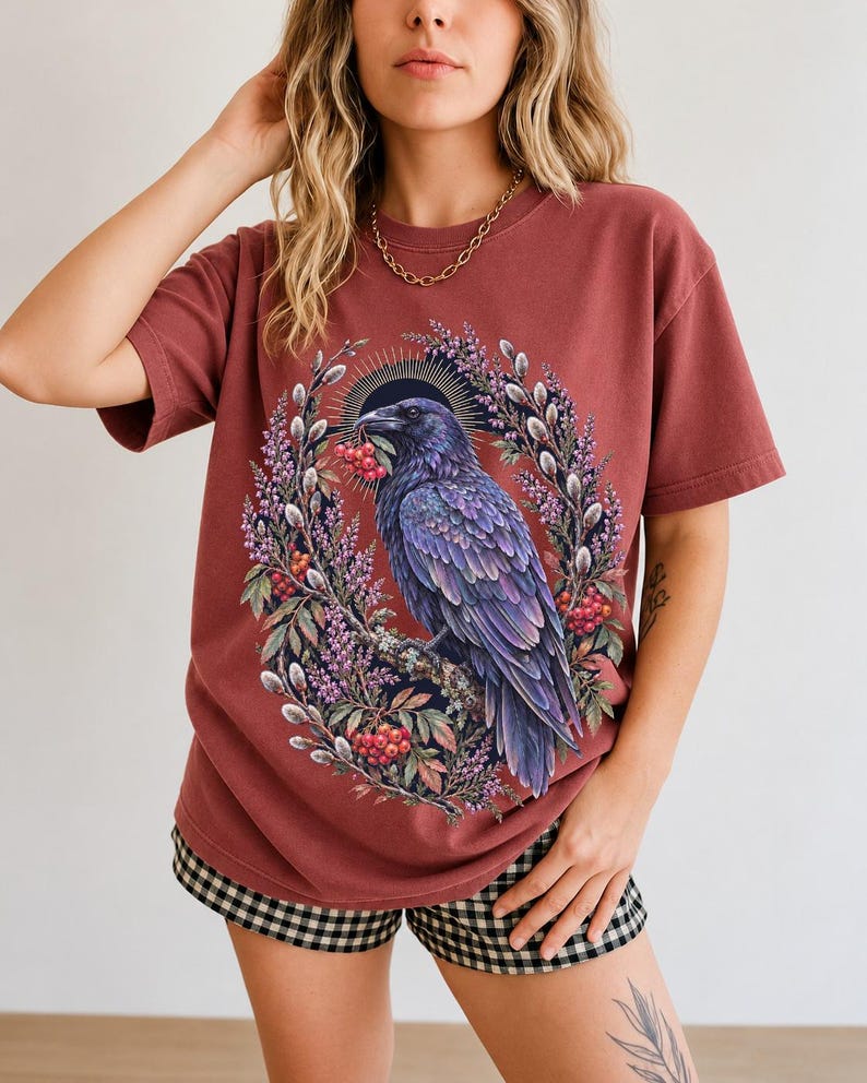 Raven holding a rowan berry inside an oval wreath of heather and catkins, painted cottagecore bird illustration