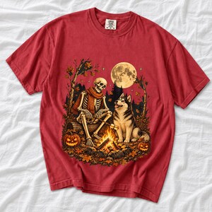 A skeleton in a scarf roasting marshmallows by a campfire beside a husky in an autumn forest under a full moon