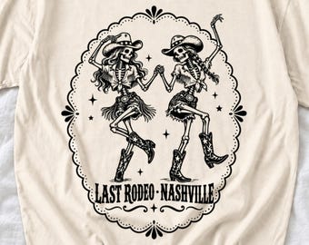 May include: A cream-colored t-shirt featuring a black graphic of two skeletons wearing cowboy hats, fringed tops, and boots, dancing within an ornate oval frame. The text "LAST RODEO - NASHVILLE" is printed in black at the bottom of the frame.