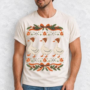Three geese in pompom knit beanies with mittens, snowflakes and pine borders, retro winter t-shirt graphic
