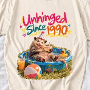 May include: A cream-colored t-shirt featuring a graphic of an opossum wearing a party hat in a blue kiddie pool. The opossum holds a slice of cake. Text reads "Unhinged Since 1990". A beach ball, a rubber duck, and a glass of orange juice are visible.